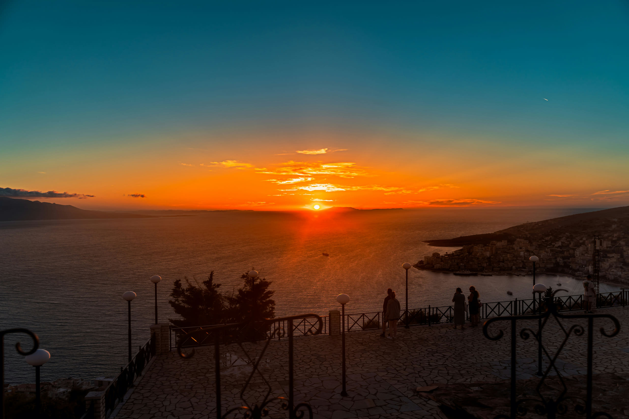 Sonnenuntergang über einer Bucht an der albanischen Küste, im Vordergrund eine Promenade mit schmiedeeisernem Geländer und Laternen